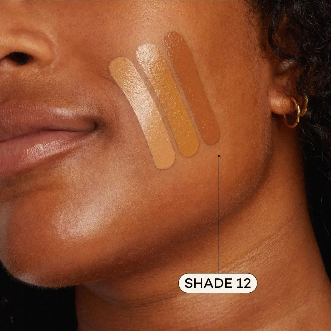 A close-up of a woman’s cheek with three Axiology Fungdation Mushroom Foundation swatches in various brown tones; the darkest shade is labeled SHADE 12 with an arrow pointing to it.