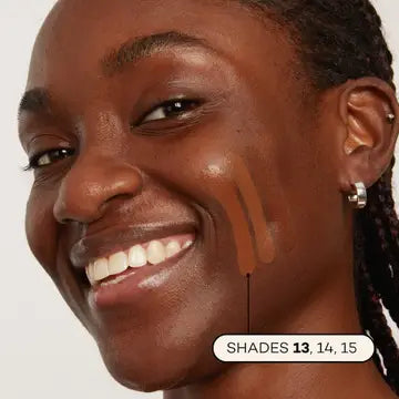 A smiling person with braided hair displays three Axiology Fungdation Mushroom Foundation swatches—shades 13, 14, and 15—on their cheek. Their glowing skin and a small hoop earring emphasize the foundation’s flawless finish.