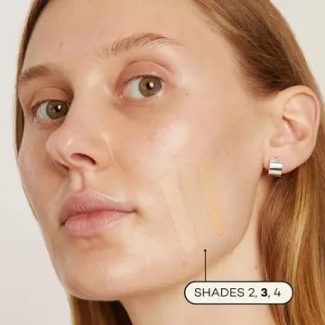 A woman with fair skin and light brown hair applies three swatches of Axiology Fungdation Mushroom Foundation in shades 2, 3, and 4 on her cheek. She wears a silver hoop earring and gazes slightly away from the camera.