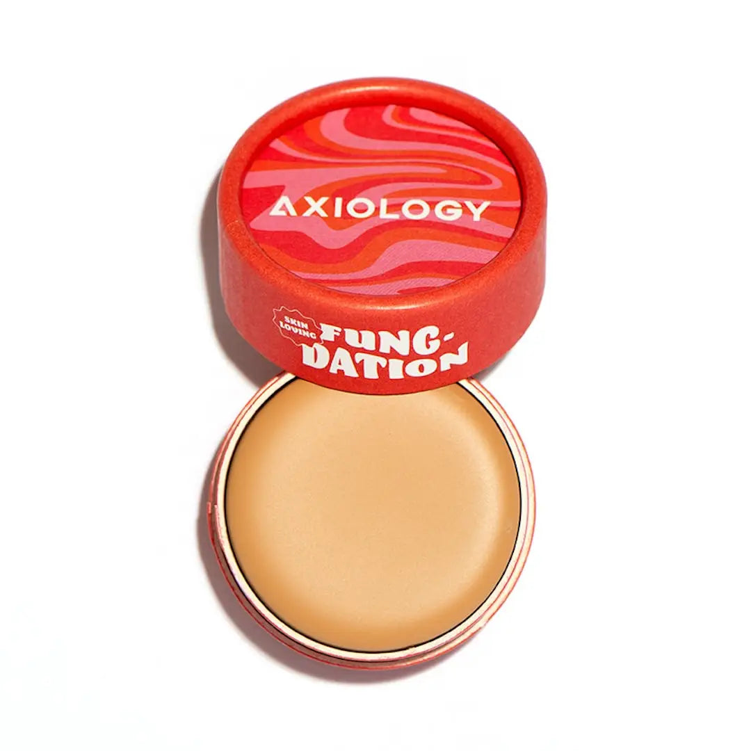 Axiology Fungdation Mushroom Foundation is shown in a round red container with the lid partially off, revealing beige cream makeup inside. The wavy-patterned lid features white text, and the hydrating foundation sits on a white background.