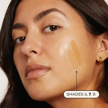 A woman with medium skin tone wears Axiology Fungdation Mushroom Foundation in shades 6, 7, and 8 swatched on her cheek. She has dark hair, minimal makeup, and small gold hoop earrings.