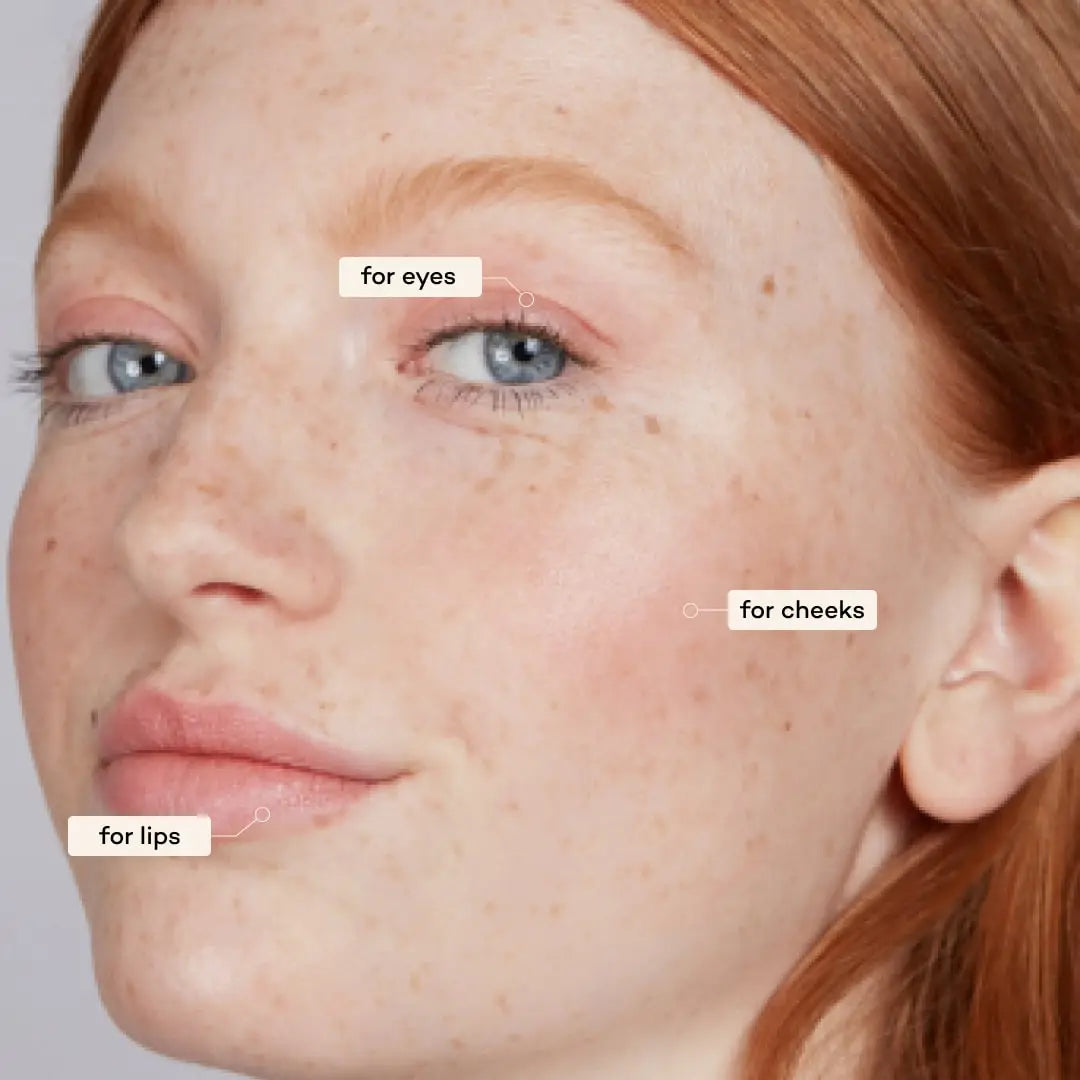 A close-up of a woman with fair skin, red hair, and blue eyes features Axiology 3-in-1 Balmie Crayon Singles applied on eyelids, cheekbones, and lips—highlighting plastic-free makeup versatility.