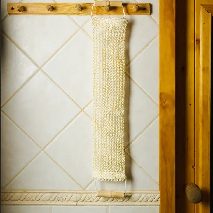 The Bamboo Switch Sisal Linen Back Exfoliating Strap, a long eco-friendly cream product, hangs from two wooden pegs on a white tiled wall with wood trim.