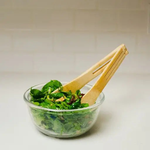 Glass bowl with green salad and wooden tongs on a white surface