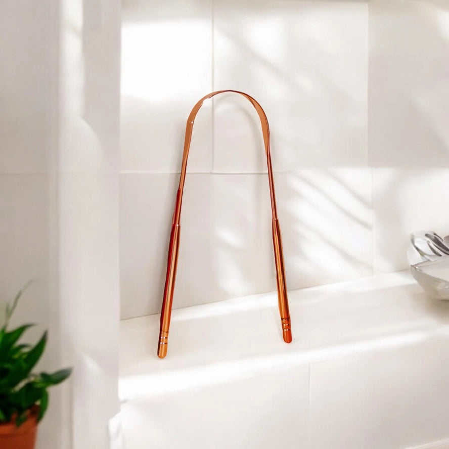 rose gold tongue scraper propped up against white bathroom tile