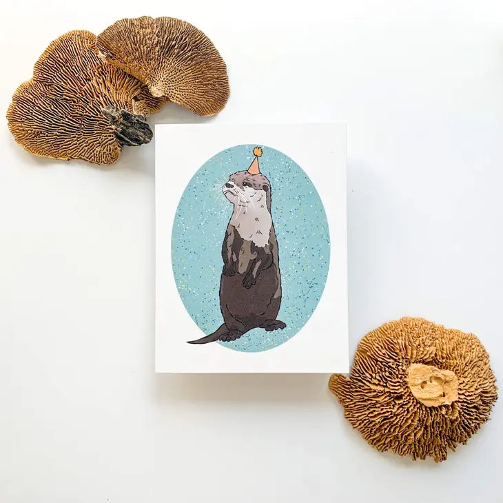 A Bean Horse Car Designs Greeting Card features an illustrated otter in a party hat on a blue speckled background, displayed on a white surface with three large, textured brown mushrooms nearby.