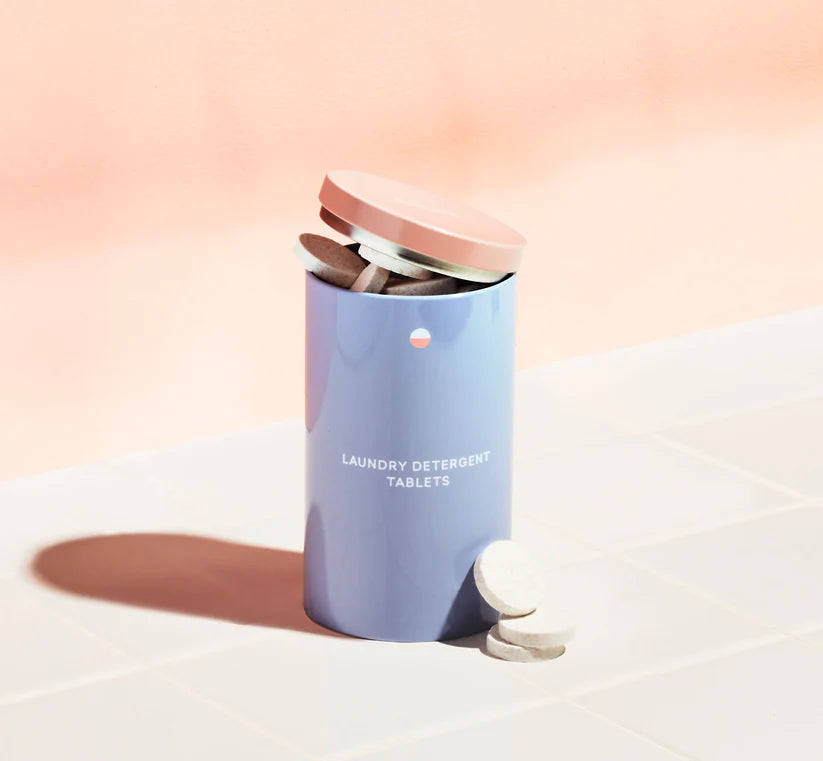 A pastel blue Blueland Laundry Tablets container with a pink lid sits open on white tiles, spilling out plastic-free tablets. The background is a soft peach color.
