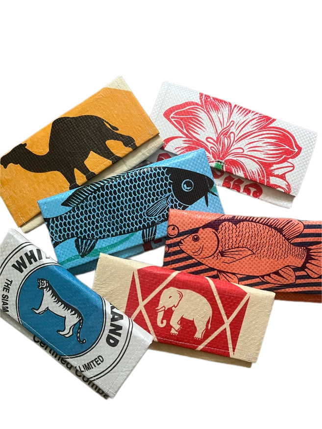 Six colorful rectangular Cement6 Accordion Clutch bags, each artisan made with bold graphic illustrations—a camel, a flower, two types of fish, a tiger, and an elephant—are thoughtfully arranged on a white background.