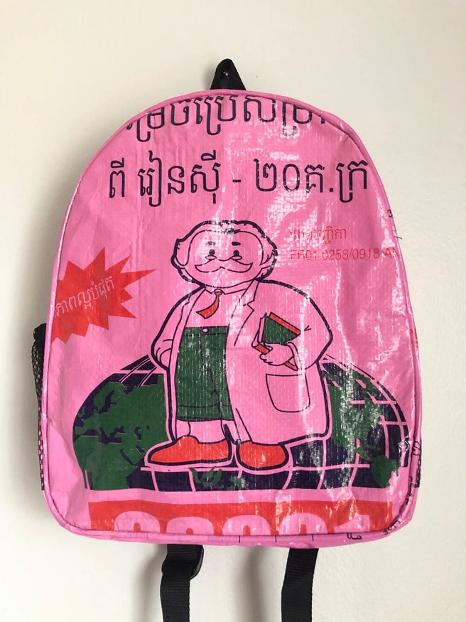 The Cement6 Minimal Daypack is a pink artisan-made backpack featuring a cartoon older man with white hair, mustache, lab coat, and green outfit holding a book, plus bold graphics and Khmer text.