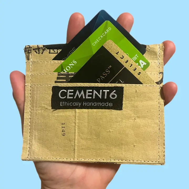 A hand holds a tan Cement6 Wallets card carrier, artisan-made and minimalistic, with four credit and debit cards partially visible against a light blue background.