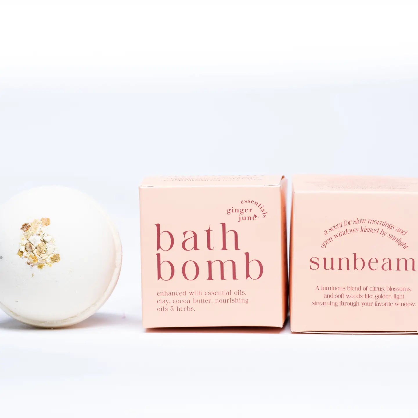 A white Ginger and June Botanical Bath Bomb with a gold leaf accent is displayed next to two pink boxes labeled "Bath Bomb" and "Sunbeam," featuring essential oils, cocoa butter, and a citrus scent blend.