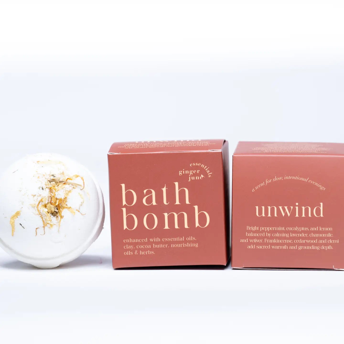 A white Botanical Bath Bomb with dried flower petals from Ginger and June rests beside two pink boxes, featuring essential oils, ginger, juniper, and soothing ingredients for a relaxing bath experience.