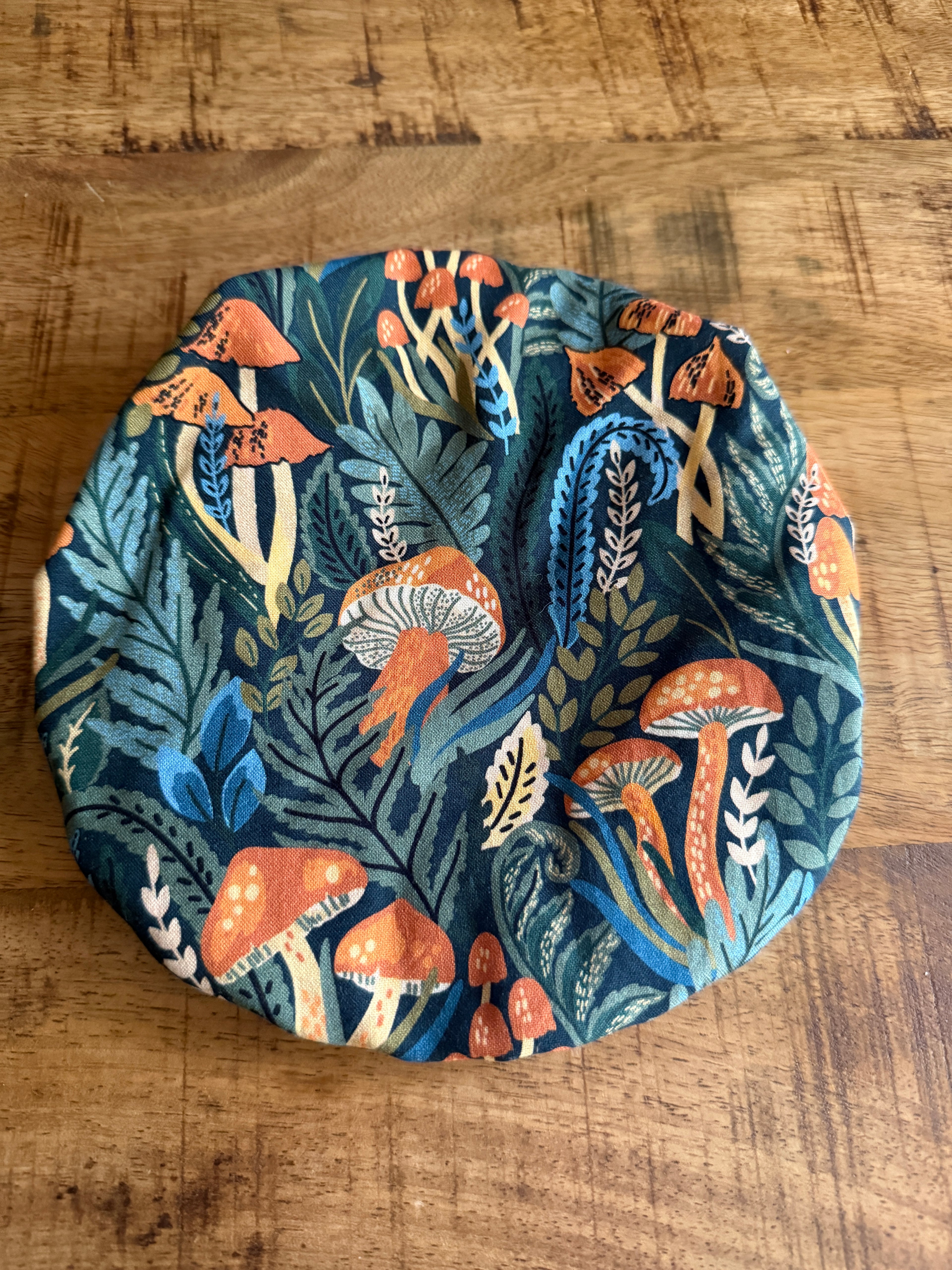 The Sustainable For Good Reusable Reversible Bowl Cover, featuring orange mushrooms, green leaves, and blue foliage, adds color to your plastic-free kitchen and is shown here on a wooden surface.
