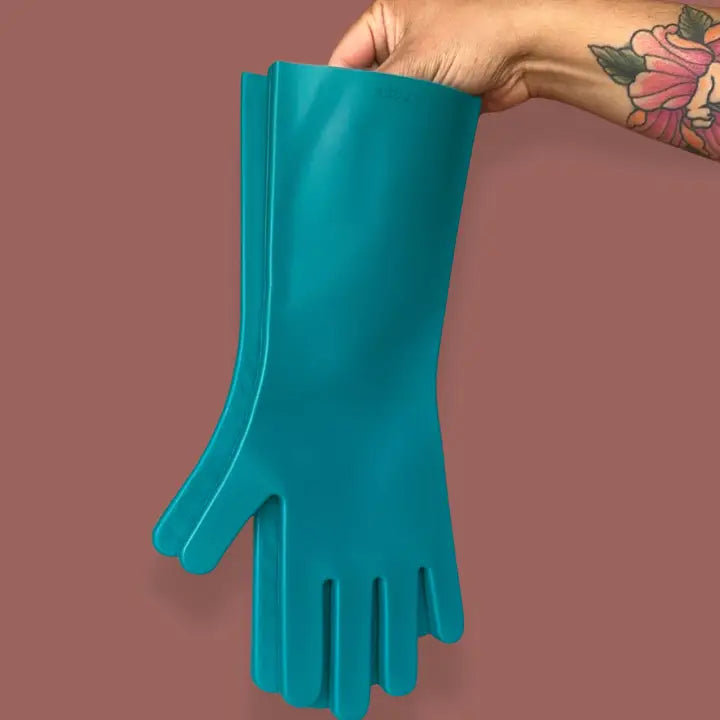 A hand with a floral tattoo holds up a pair of Keeps Silicone Cleaning Gloves in teal against a plain mauve background.
