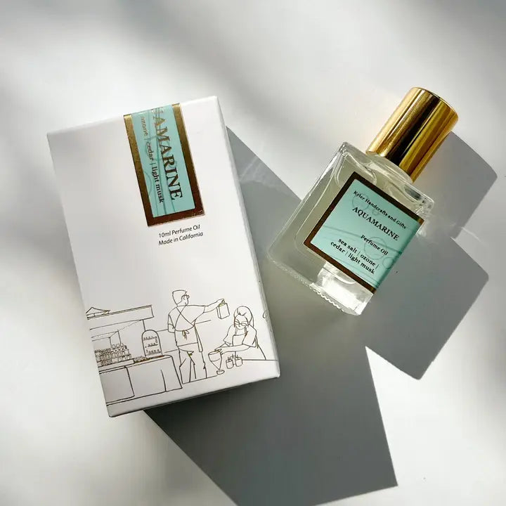 A Kyler Handcrafts Roll On Perfume with a gold cap rests by its illustrated white box with gold details, bathed in soft natural light. Phthalate-free, this handcrafted fragrance exudes elegance and charm.
