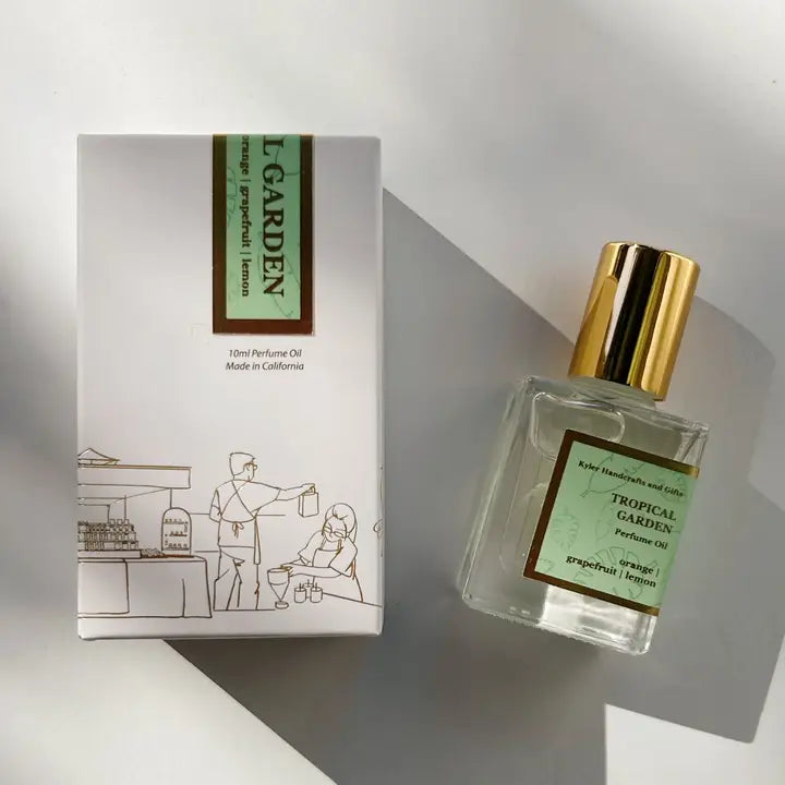 A bottle of Kyler Handcrafts Roll On Perfume with a gold cap sits beside its white box, decorated with a minimalist line drawing of two people at a market stall. Sunlight casts gentle shadows on this handcrafted scent.