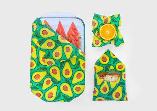 Three foods—a sandwich, watermelon slices in a container, and a halved orange—are wrapped in Meli Wraps Beeswax Wraps (3 pk) with an avocado design, highlighting stylish eco-friendly storage against a plain light background.