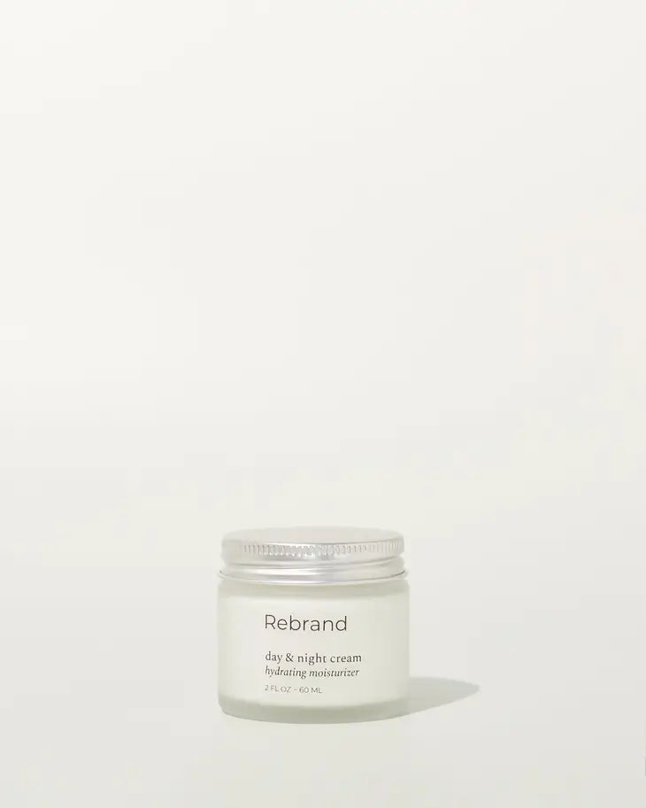 A small, clear jar with a silver lid labeled Rebrand Day & Night Cream rests on a light background—your sustainable, hyaluronic acid moisturizer in a refillable design.