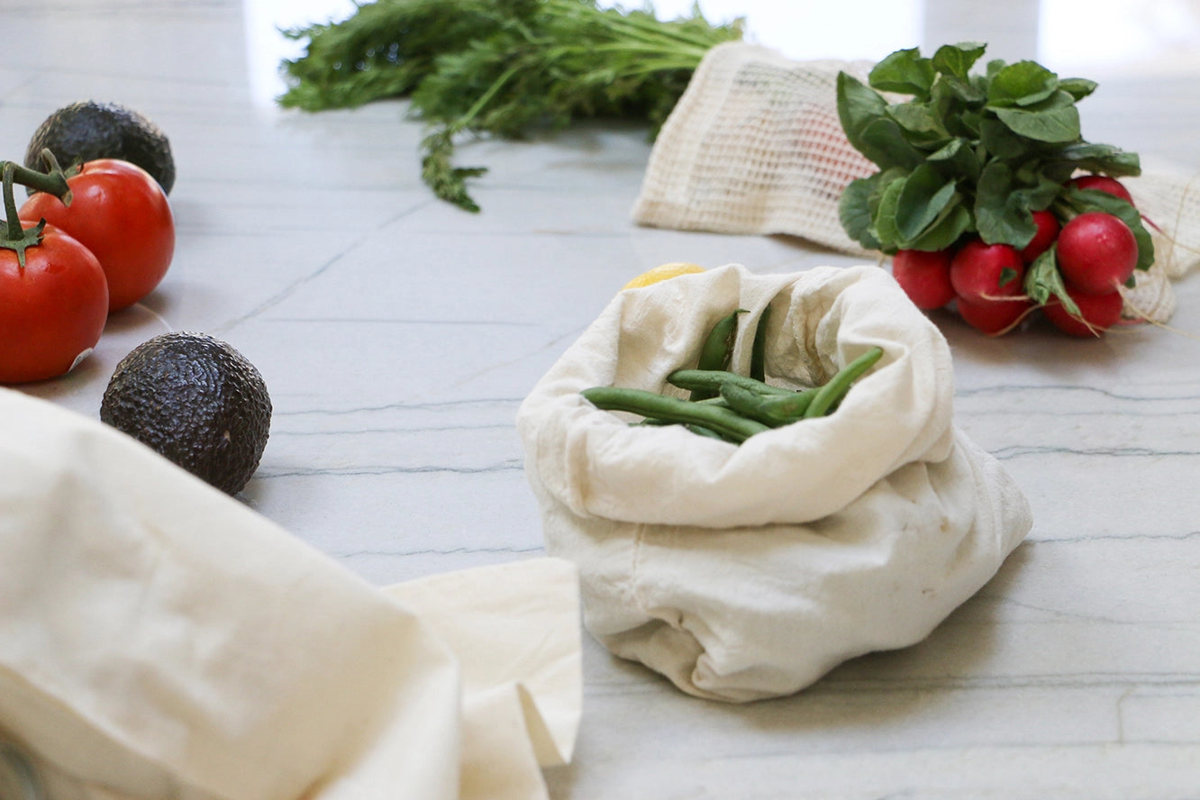 muslin produce bags with green beans in it, tomatoes, avocado and radishes to the side
