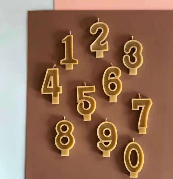 Ten Beeswax Number Candles by The Sud Sister, shaped 0-9, stand upright with visible wicks on a brown and pink background. Made from sustainable beeswax, each candle clearly displays its number.