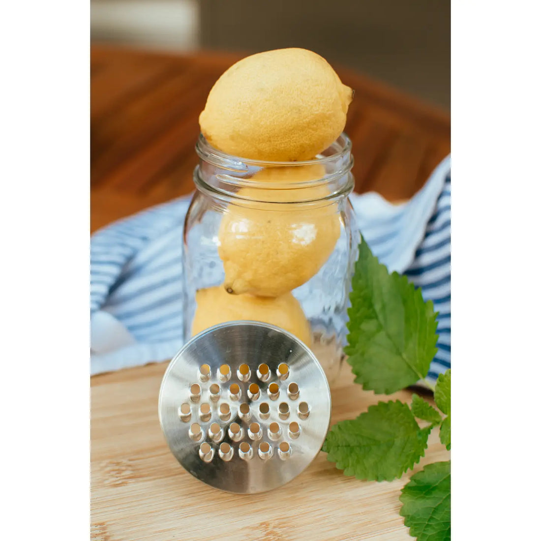 Glass jar with lemons, metal strainer, and green leaves on a wooden surface.
