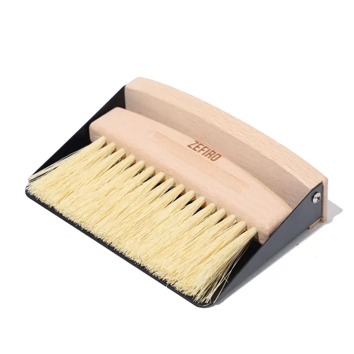 The Zefiro Mini Sweep features a mini broom with yellow bristles and a beechwood handle, labeled "ZEFIRO," resting inside a black metal dustpan. Both compostable items are displayed on a white surface.
