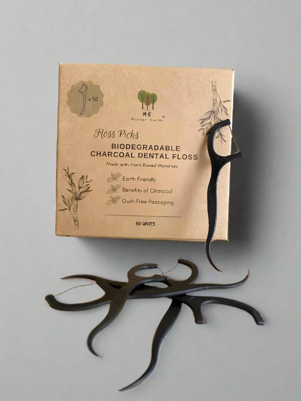 A box labeled "Me.Mother Earth Biodegradable Charcoal Dental Floss Picks" with black bamboo charcoal floss picks scattered in front. The packaging emphasizes eco-friendly, plant-based, and plastic-free dental picks.