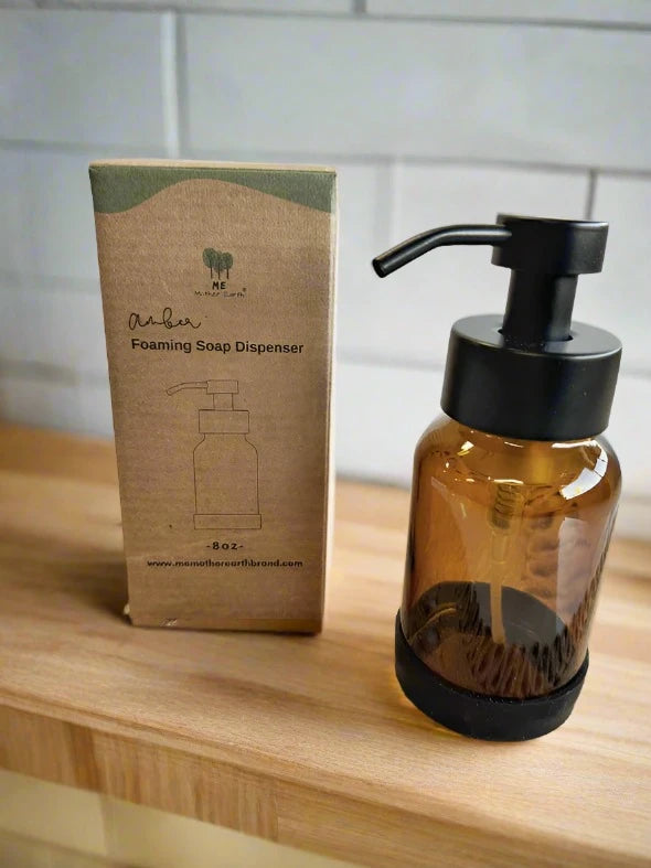 The Me.Mother Earth Amber Foaming Soap Dispenser with a black pump stands beside its cardboard box labeled "Amber Foaming Soap Dispenser" and displays a simple line drawing of the product.