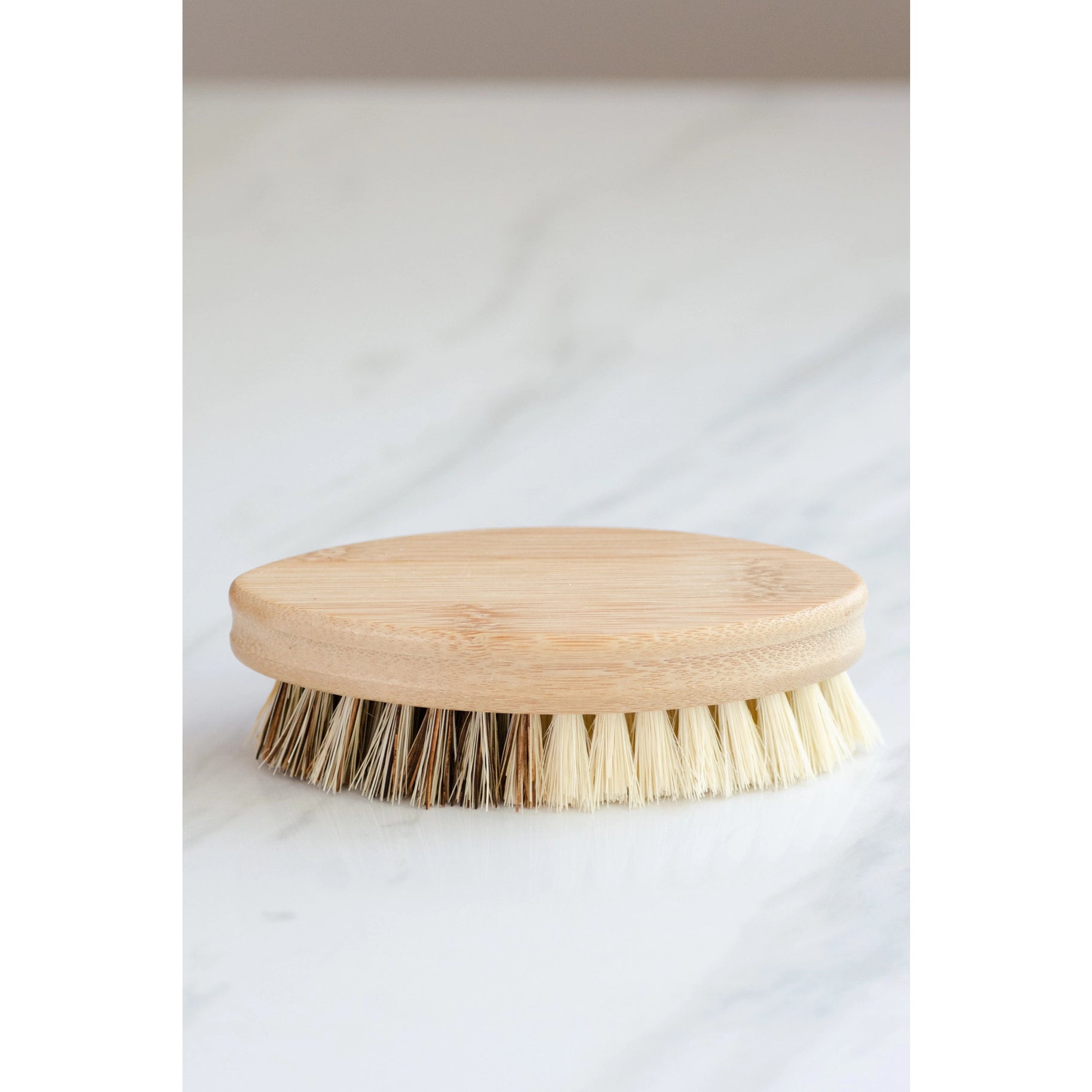 The No Tox Life Vegetable Brush, featuring a round bamboo handle and light bristles, rests on a white marble surface.