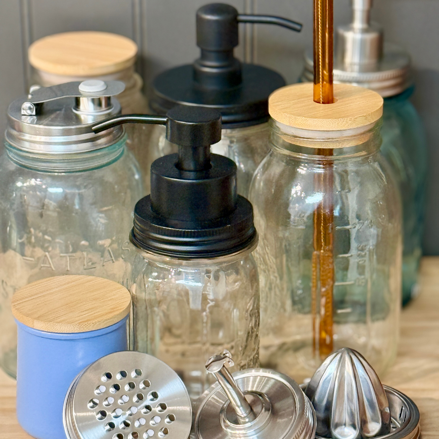 A group of empty glass jars, some with metal or bamboo lids, pumps, or straws. In front are various metal kitchen tools, including a juicer, dispenser, and shaker lid.