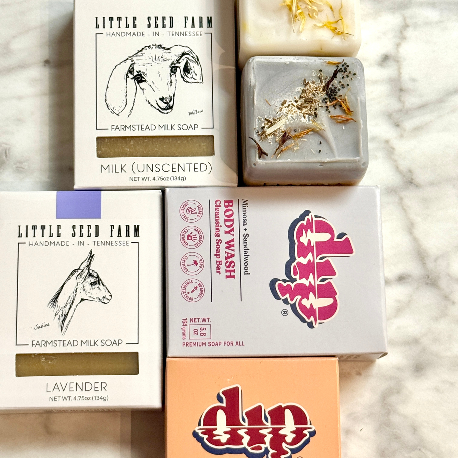 Five artisan soap bars, some in boxes, are arranged on a marble surface. The boxes feature illustrated labels, including goat images and Little Seed Farm. Two soap bars are unboxed, showing textured tops.