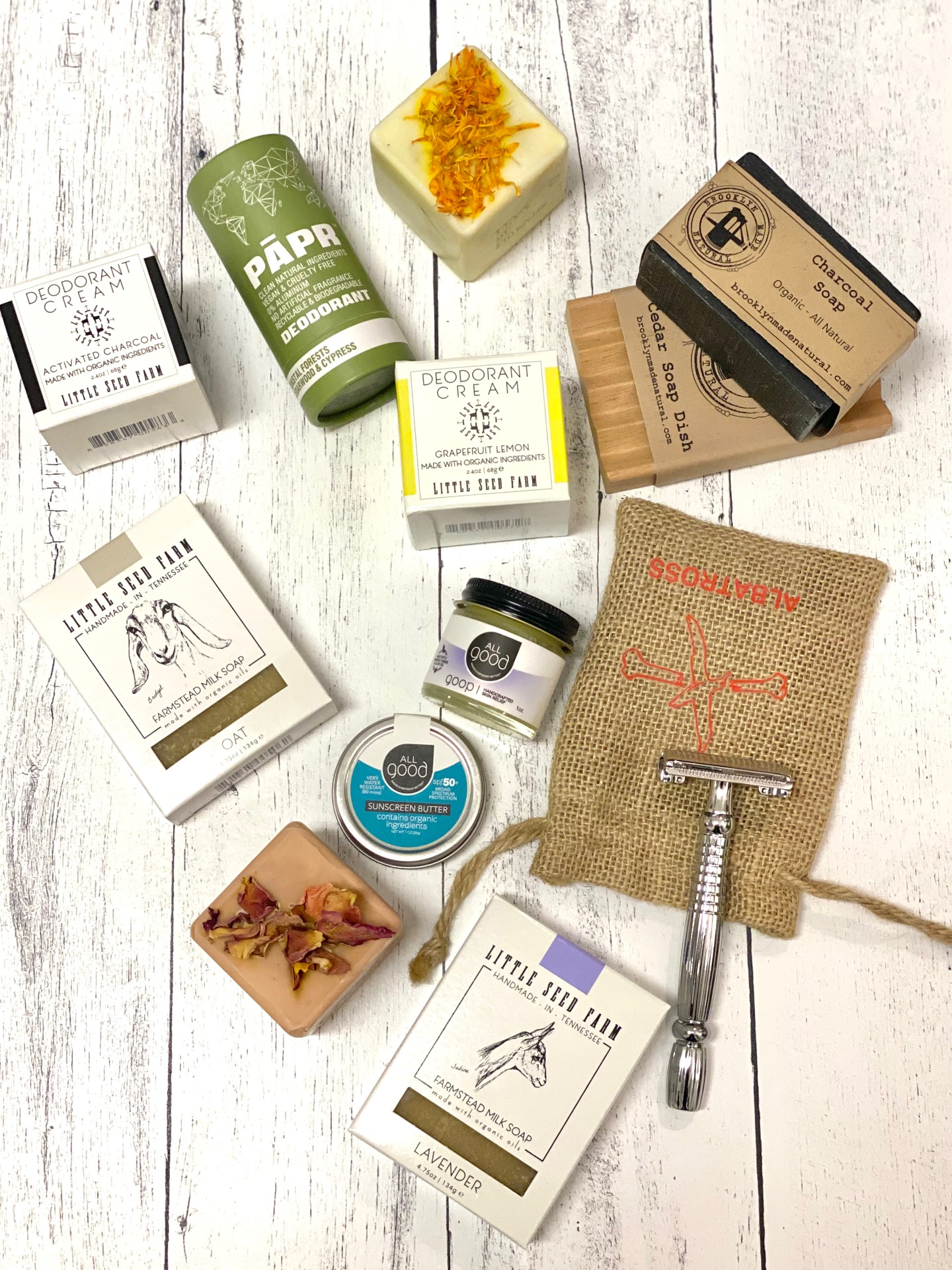 A flat lay of eco-friendly personal care products, including deodorants, soap bars, lotion, a safety razor, and a burlap pouch, all arranged on a white wooden surface.