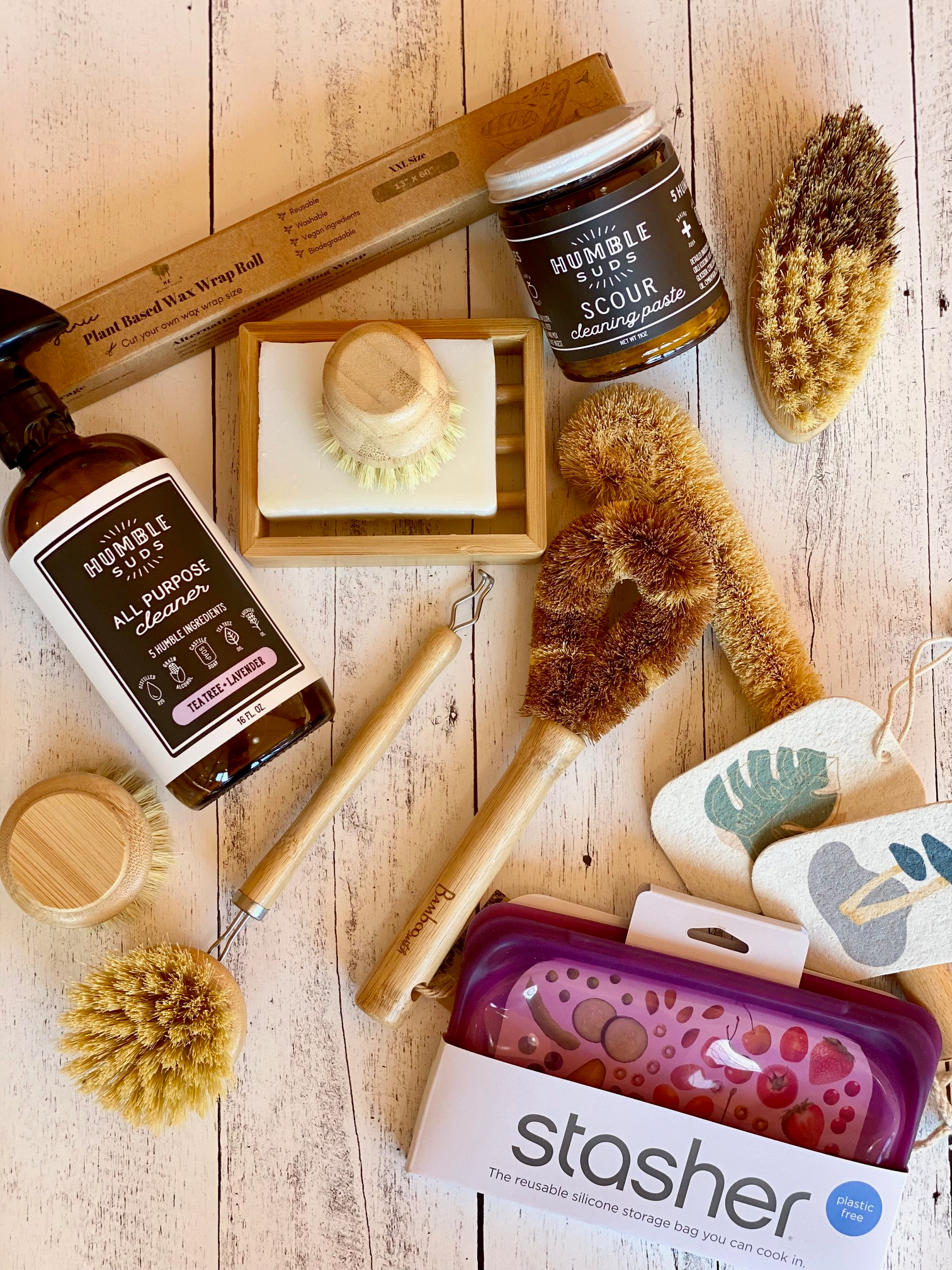 A flat lay of eco-friendly cleaning products, including brushes, a spray bottle, cleaning paste, a bamboo toothbrush, a reusable silicone storage bag, and natural sponges on a white wooden surface.