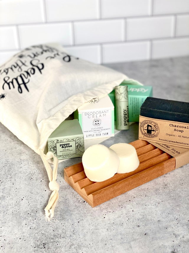 A cloth bag with text spills out natural skincare products, including green boxes, a charcoal soap bar, and two round soaps on a wooden tray, all on a light gray surface.