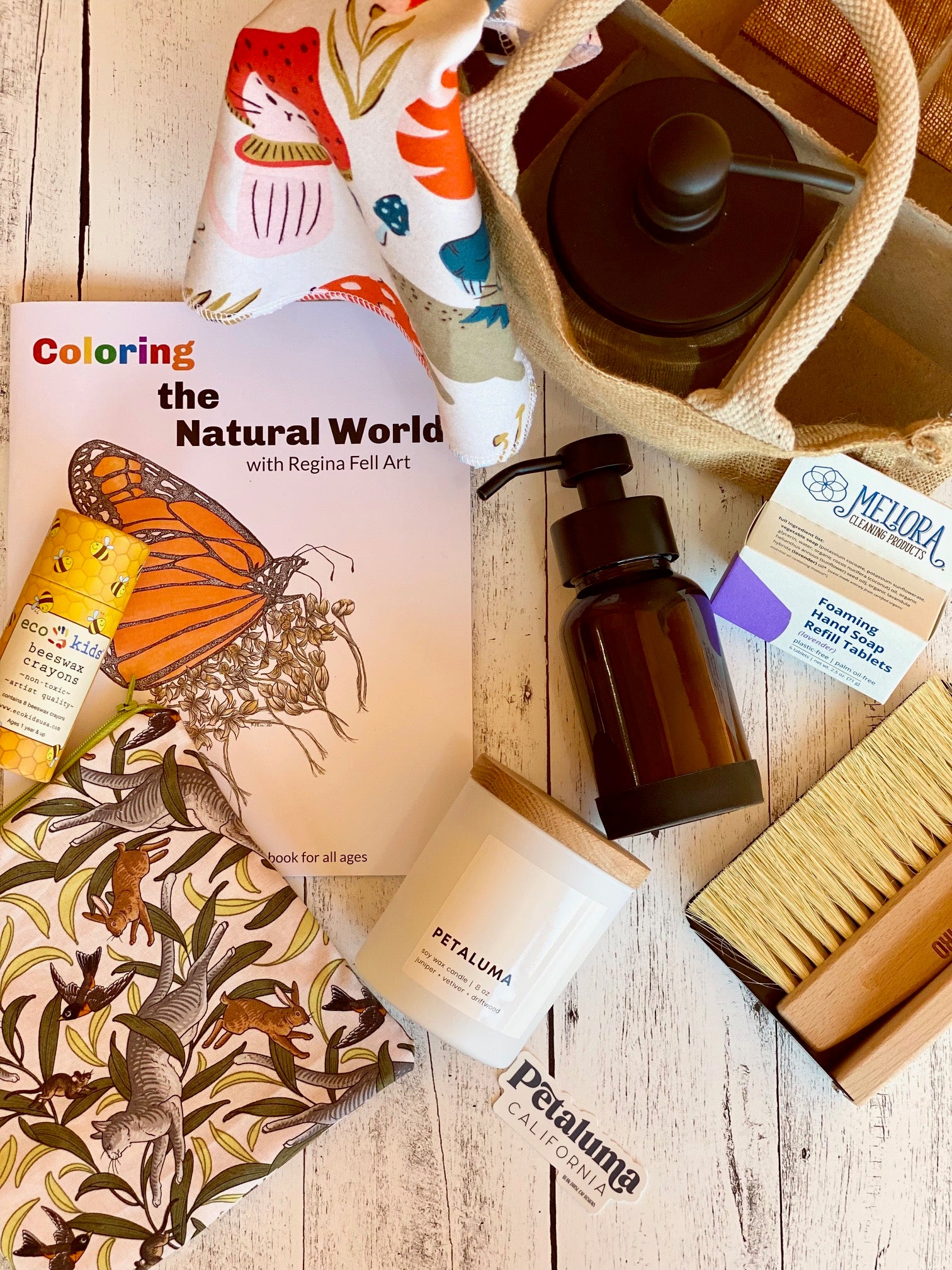A flat lay of eco-friendly products including a coloring book with a butterfly cover, a reusable bag, candle, soap bar, lotion, cleaning brush, and fabric items on a white wooden surface.