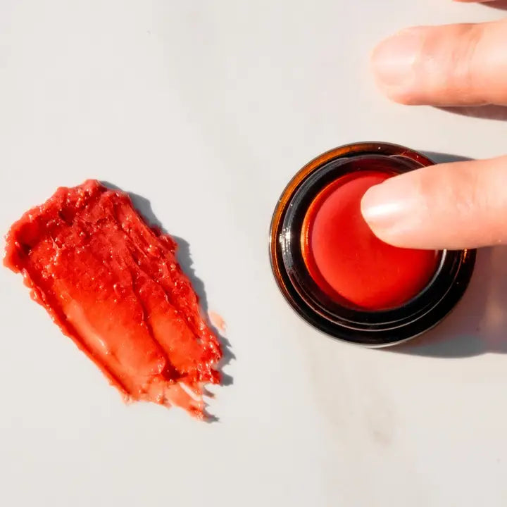 A hand holds an open tin of 8 Days Botanicals Rose Petal Lip Butter, with a smear of the natural tinted moisturizer displayed on a nearby light surface.