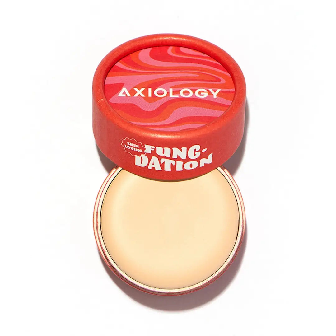 An open round container of Axiology Fungdation Mushroom Foundation with a red and pink patterned lid above it reveals smooth beige foundation inside. The lid features the Axiology logo and product name. 