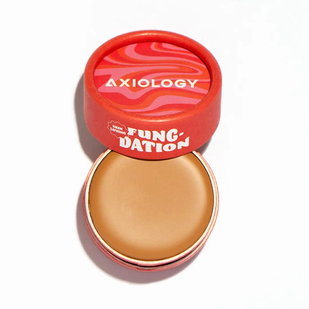 An open Axiology Fungdation Mushroom Foundation reveals tan cream inside its round container. The red lid, with wavy patterns and bold white and yellow branding, hints at its hydrating satin finish formula.