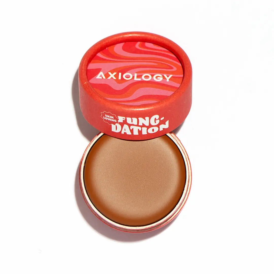 A round container of Axiology’s Fungdation Mushroom Foundation, with the lid partially open to reveal the smooth, brown makeup. The red packaging features a wavy pink and orange design on the lid.