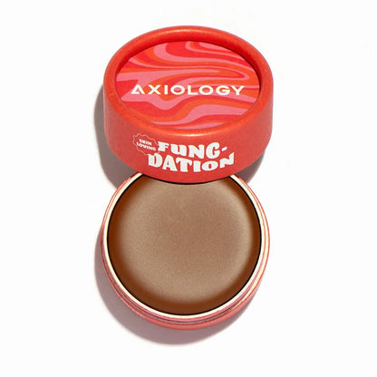 A round red container with a swirl-patterned lid labeled Axiology holds Fungdation Mushroom Foundation. The lid is partially off, revealing the smooth, hydrating brown foundation inside.