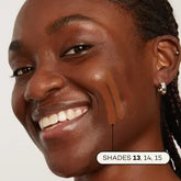 A smiling person with braided hair displays three Axiology Fungdation Mushroom Foundation swatches—shades 13, 14, and 15—on their cheek. Their glowing skin and a small hoop earring emphasize the foundation’s flawless finish.