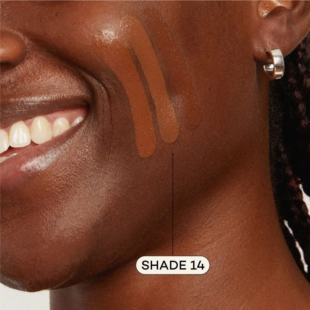 Close-up of a smiling person with dark skin, silver hoop earring, and three Axiology Fungdation Mushroom Foundation swatches on their cheek. An arrow points to the most blended swatch labeled SHADE 14.