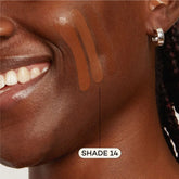Close-up of a smiling person with dark skin, silver hoop earring, and three Axiology Fungdation Mushroom Foundation swatches on their cheek. An arrow points to the most blended swatch labeled SHADE 14.