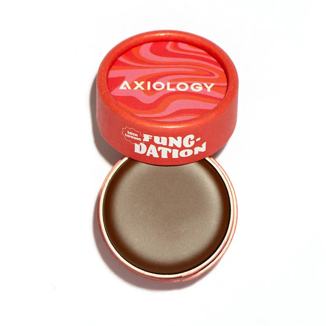 An open, round red container labeled "Axiology Fungdation Mushroom Foundation" with a swirled pink-red lid reveals a neutral-toned cream foundation inside.