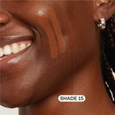 A close-up shows a smiling person’s cheek with three tones of Axiology Fungdation Mushroom Foundation swatched. SHADE 15 is labeled on the bottom swatch. The person wears a silver hoop earring.