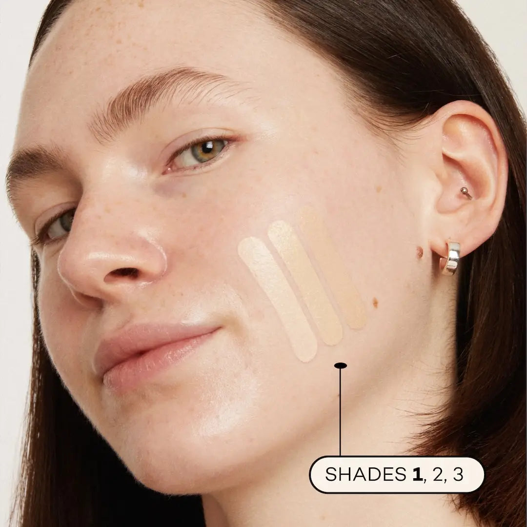 A woman with fair skin and brown hair wears silver hoop earrings. Three swatches of Axiology Fungdation Mushroom Foundation, labeled shades 1, 2, and 3, are applied to her cheek. 