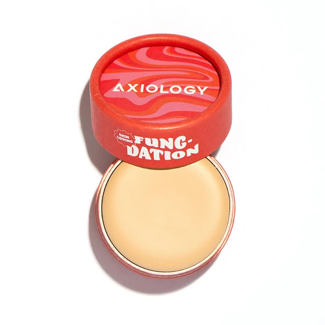 An open Axiology Fungdation Mushroom Foundation shows its light beige, hydrating cream in a round container with a red, wavy-patterned lid, elegantly displayed on a white background.