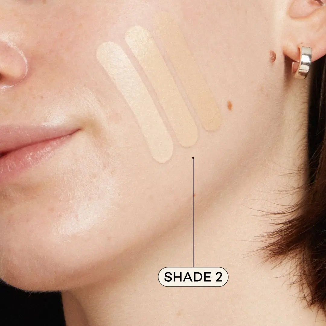 A close-up of fair skin with a beauty mark and a small hoop earring displays three swatches of Axiology Fungdation Mushroom Foundation. An arrow highlights the middle swatch, labeled Shade 2. 