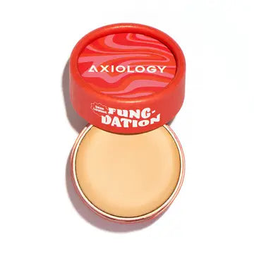A round, red container labeled "Axiology Fungdation Mushroom Foundation" sits with its wavy red and pink lid partially open, showing the beige satin-finish foundation inside, enriched with gentle mushroom ingredients.