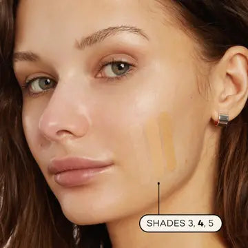 A woman with light skin and green eyes wears minimal makeup and a silver hoop earring. She has three swatches of Axiology Fungdation Mushroom Foundation in shades 3, 4, and 5 on her cheek.